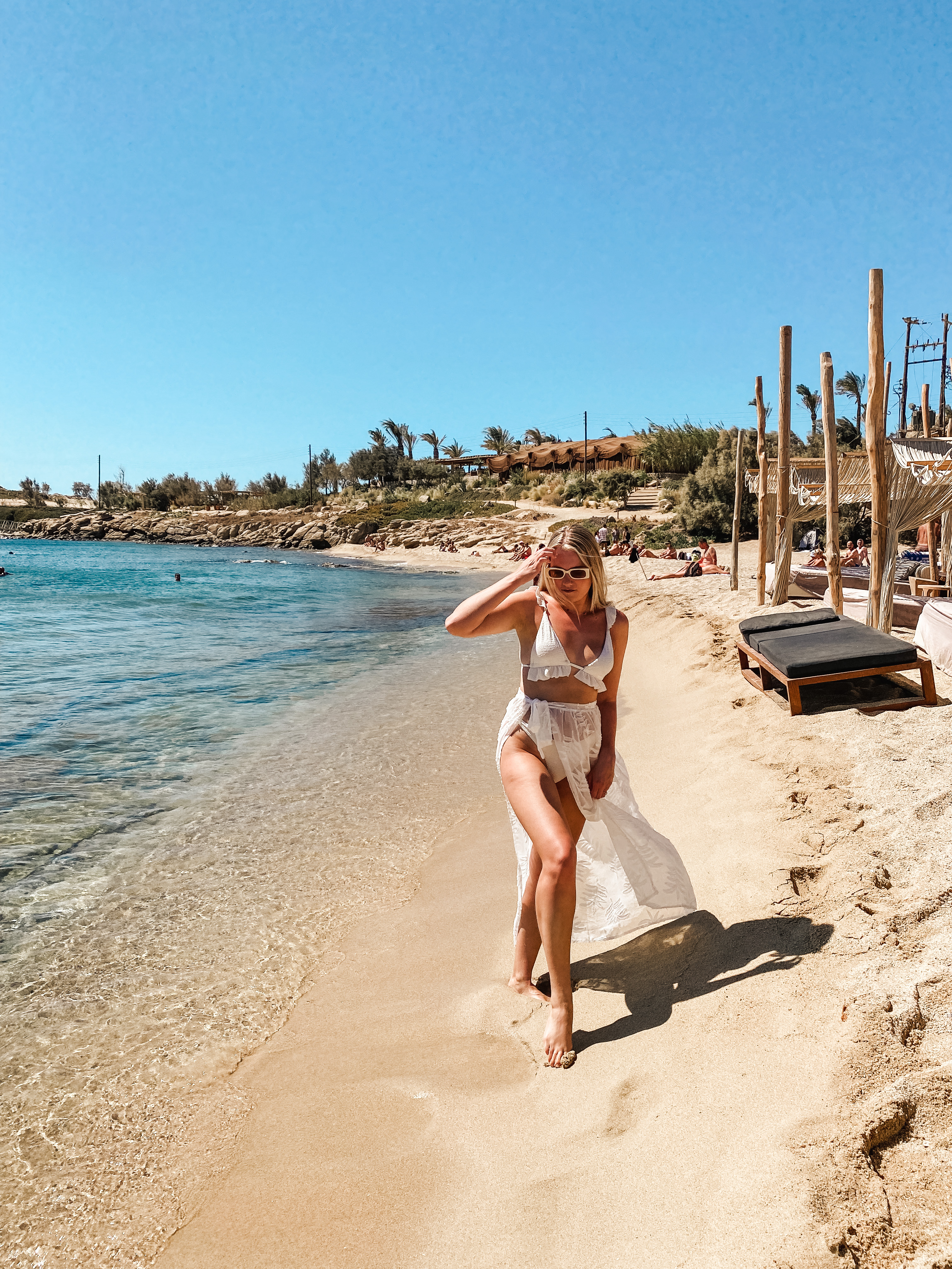 Blonde lounging in white bikini walking along Mykonos Greece Beach