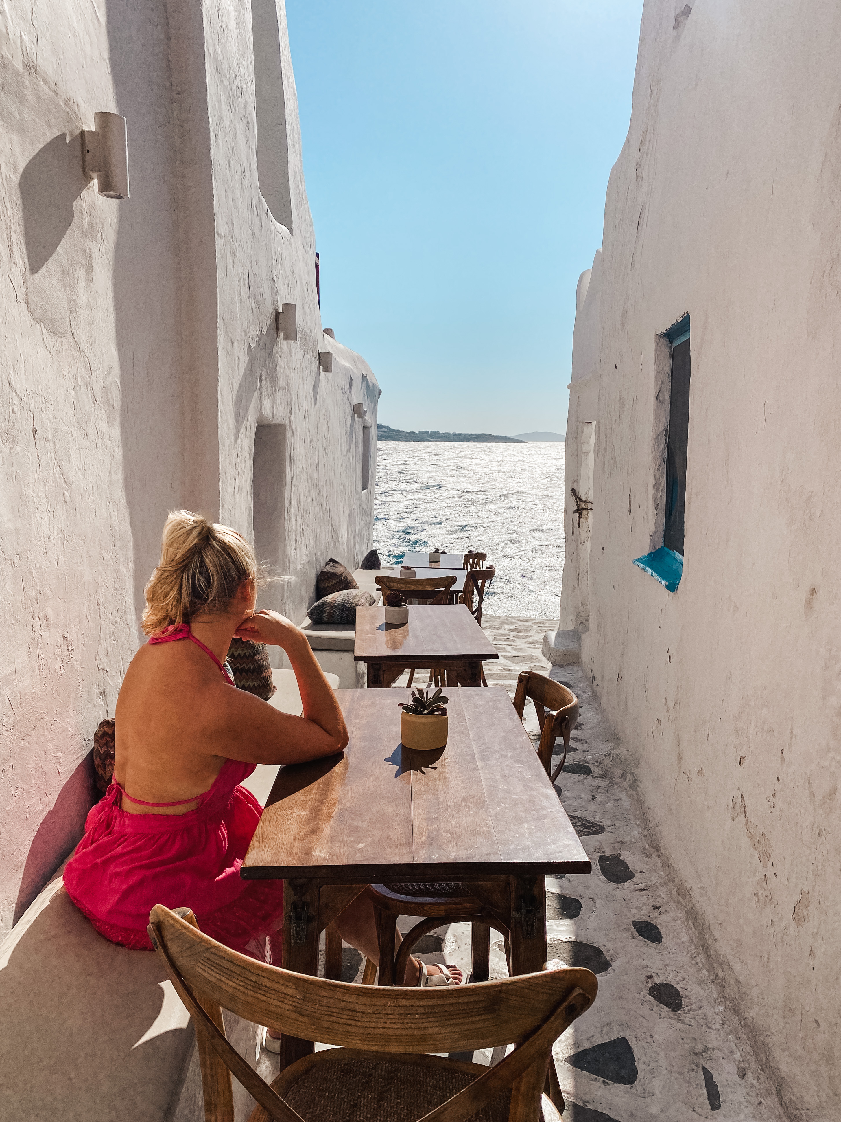 Blonde in pink dress sitting at a tabel in white washed alley, Mykonos Greece