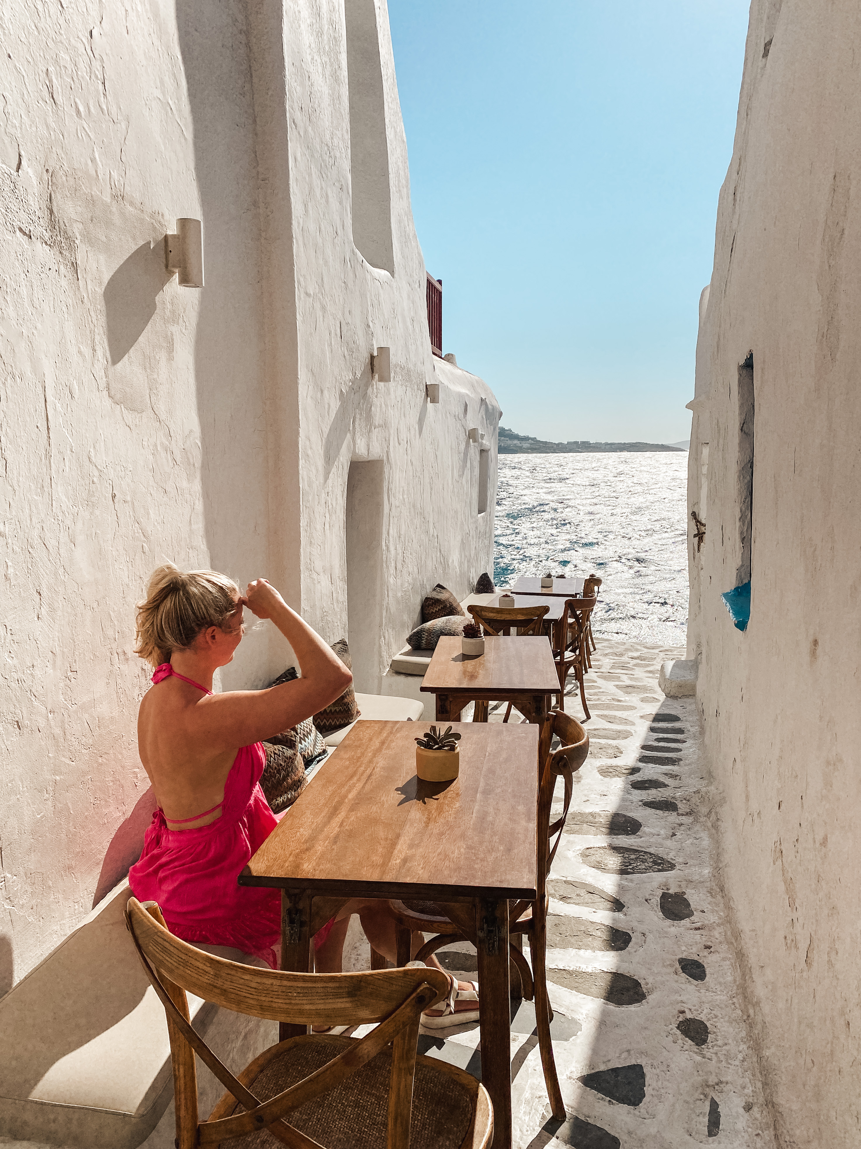 Blonde in pink dress sitting at a tabel in white washed alley, Mykonos Greece